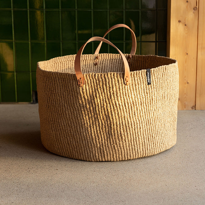 Woven basket with leather handles on a concrete surface near a glass door.