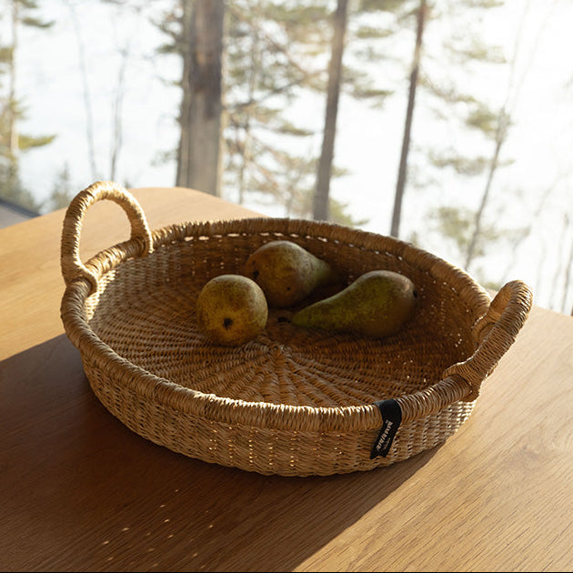 Woven basket with fruits on a wooden surface near a window with a view of trees.
