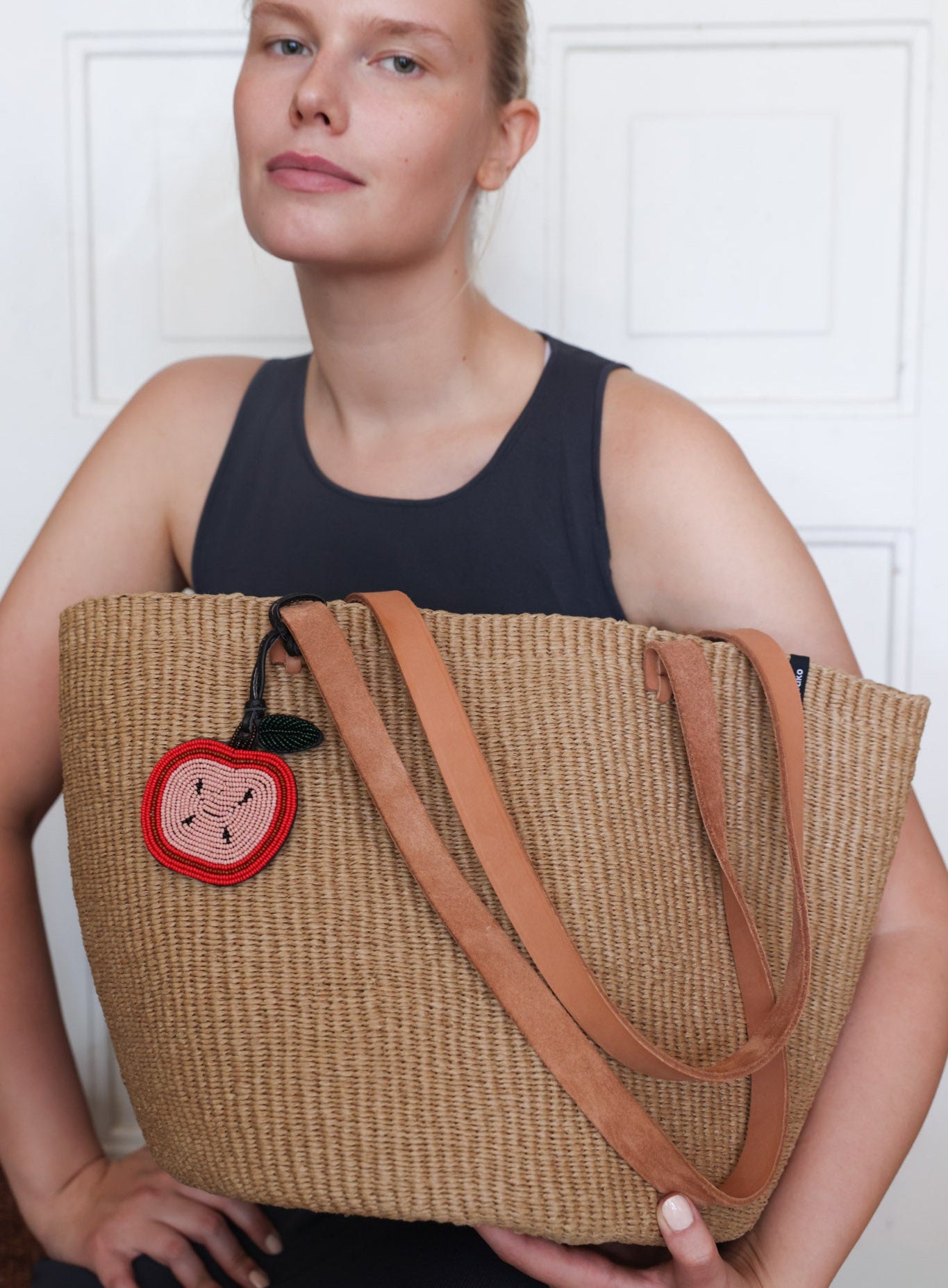 A woman wearing a black tank top holds a large woven straw tote bag with tan leather shoulder straps and a red embroidered apple patch on the front, set against a white paneled wall.
