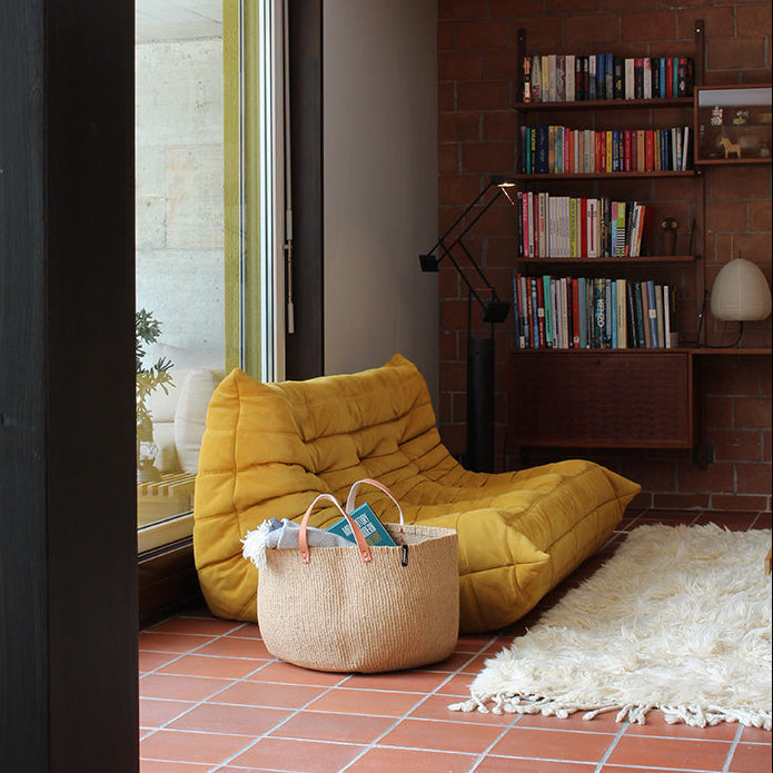 Living room with yellow sofa, handwoven brown basket, bookshelf, and brick wall.
