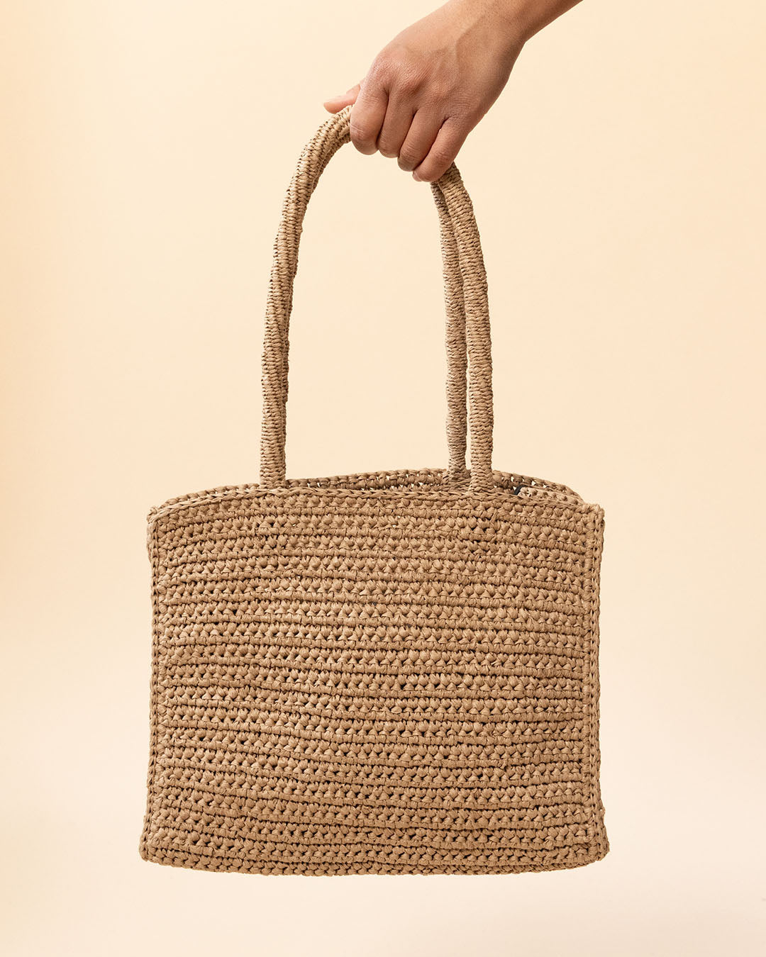 Hand holding a woven brown bag against a beige background